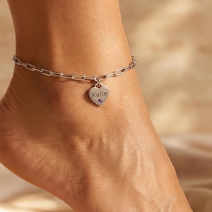 Peut inclure: Une chevillère argentée composée d'une fine chaîne à maillons et d'un petit pendentif en forme de cœur. Le pendentif porte l'inscription gravée Katie et est orné d'une minuscule pierre décorative foncée incrustée à la base du cœur.
