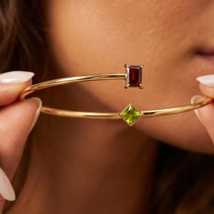 Puede incluir: Una pulsera abierta de oro con dos piedras preciosas distintas. Un extremo tiene una piedra granate rectangular de color rojo intenso, mientras que el otro extremo presenta una piedra peridoto cuadrada de color verde claro. La banda es fina y dorada.