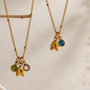 May include: Two gold necklaces, each with an 'A' pendant and gemstones. One features a blue gem, the other green and clear gems. The necklaces hang from a branch, set against a neutral backdrop. The chains are delicate.