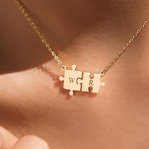 May include: A gold-toned necklace featuring two interlocking puzzle piece pendants. Each puzzle piece is engraved with a single letter, specifically the letters W and R, and the pendant is suspended from a delicate, thin gold-toned chain.