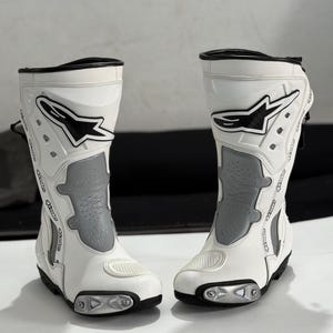 Puede incluir: Par de botas de moto blancas y grises. Las botas tienen un diseño alto, con un exterior blanco y detalles grises. Las botas presentan un logotipo negro en el lateral y están diseñadas para la protección y el rendimiento.