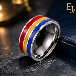 May include: A wide silver-toned metal ring featuring three horizontal enamel stripes in red, yellow, and blue. The band has a polished finish and a smooth, rounded interior. The letters EJ are visible in the upper right corner of the image.