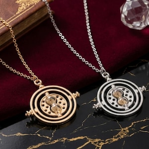 Op de afbeelding: Twee Time Turner kettingen, één goudkleurig en één zilverkleurig, elk met een ketting. De Time Turners hebben een centraal zandloperontwerp in concentrische cirkels. De kettingen liggen op een zwart-gouden oppervlak.