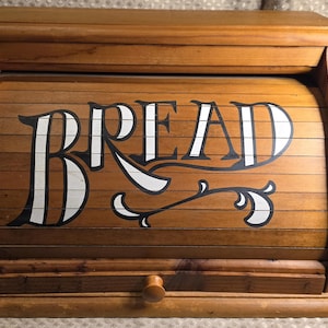 May include: A medium-brown wooden bread box featuring a roll-top door with the word BREAD printed in large, stylized white letters with black outlines. The box has a flat top, a rectangular base, and a small wooden knob on the front panel.