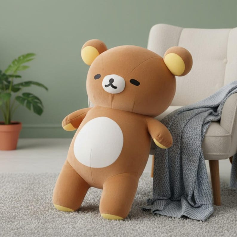 Puede incluir: Un oso de peluche marr&oacute;n con una barriga blanca, ojos negros y una peque&ntilde;a nariz negra. El oso tiene detalles amarillos en las orejas y los pies. Est&aacute; apoyado en una silla blanca con una manta gris.