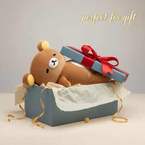 Puede incluir: Un juguete de oso de peluche marr&oacute;n con detalles amarillos, dentro de una caja de regalo azul con una cinta roja. La caja est&aacute; forrada con papel de seda blanco y cinta dorada. El texto "perfect for gift" est&aacute; encima de la caja.