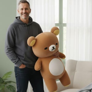 Puede incluir: Un gran oso de peluche marr&oacute;n con vientre blanco, orejas amarillas y cara en blanco y negro. El oso est&aacute; siendo sujetado por una persona que lleva una sudadera con capucha gris y vaqueros azul oscuro. El oso mide unos 61 cm de alto.
