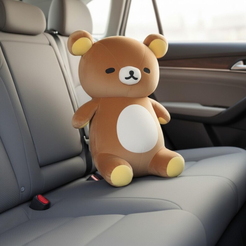 Puede incluir: Un oso de peluche marr&oacute;n con una barriga blanca y detalles amarillos en las orejas y los pies. El oso tiene nariz y ojos negros y est&aacute; sentado en un asiento de coche gris. Un juguete blando.