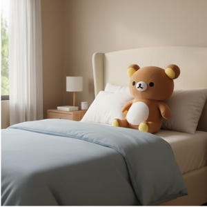 Puede incluir: Un oso de peluche marr&oacute;n con vientre blanco y detalles amarillos est&aacute; sentado en una cama con una funda n&oacute;rdica azul claro y almohadas blancas. El dormitorio tiene una paleta de colores neutros.