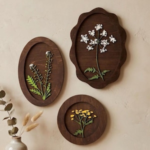 May include: Three dark brown wooden wall plaques featuring pressed botanical specimens. One oval plaque displays white flowers, a scalloped oval plaque holds a white flowering plant, and a circular plaque features a cluster of small yellow flowers.