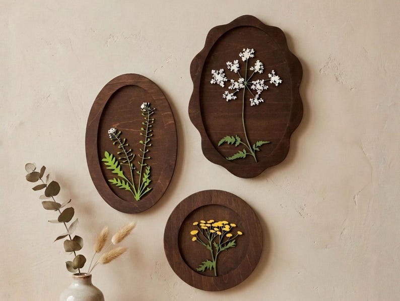 May include: Three dark brown wooden wall plaques featuring pressed botanical specimens. One oval plaque displays white flowers, a scalloped oval plaque holds a white flowering plant, and a circular plaque features a cluster of small yellow flowers.