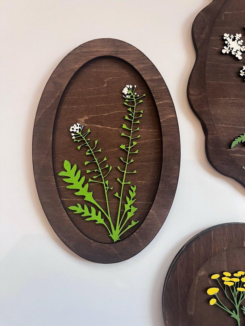 May include: An oval-shaped dark brown wooden wall plaque featuring a laser-cut green plant with small white flowers. The plant is centered within the recessed oval frame, showcasing detailed leaves and stems against the smooth wood grain background.