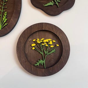 May include: A circular dark brown wooden wall plaque featuring a layered cutout of a green stem with bright yellow flower clusters. The design is recessed within the circular frame, showcasing a natural wood grain finish and precise laser-cut details.