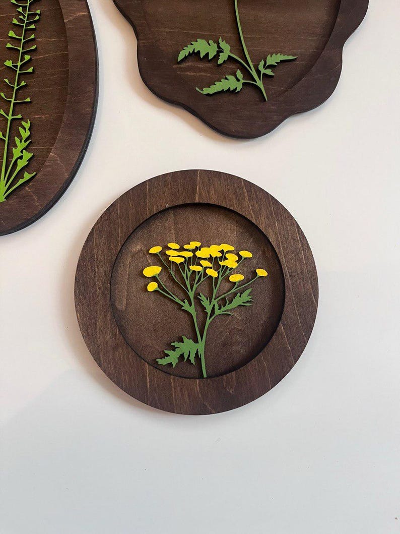 May include: A circular dark brown wooden wall plaque featuring a layered cutout of a green stem with bright yellow flower clusters. The design is recessed within the circular frame, showcasing a natural wood grain finish and precise laser-cut details.