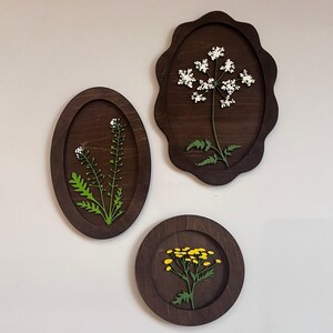 May include: Three dark brown wooden wall plaques featuring botanical cutouts. One oval plaque displays a green plant with white flowers, one scalloped plaque shows a white flower cluster, and one circular plaque features a yellow flower cluster.