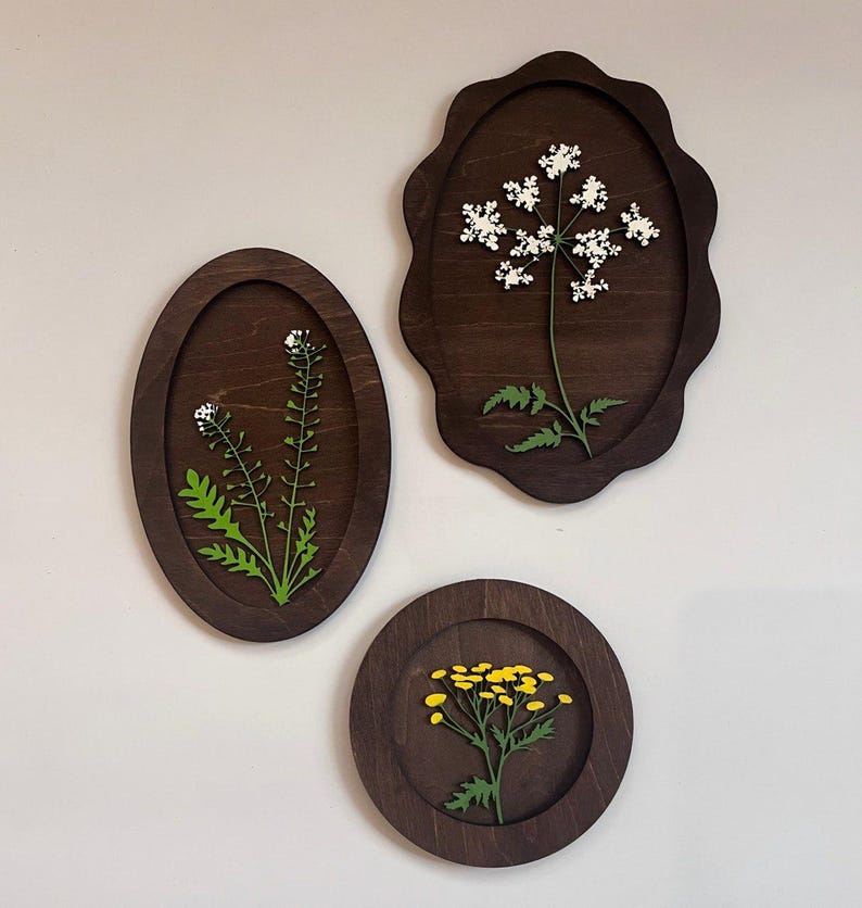 May include: Three dark brown wooden wall plaques featuring botanical cutouts. One oval plaque displays a green plant with white flowers, one scalloped plaque shows a white flower cluster, and one circular plaque features a yellow flower cluster.