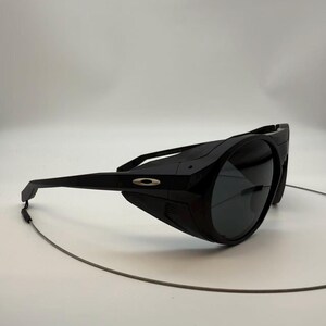Puede incluir: Gafas de sol negras envolventes con lentes oscuras y montura negra. Las gafas de sol presentan un logotipo plateado en el lateral y un fino cord&oacute;n gris sujeto a la montura.