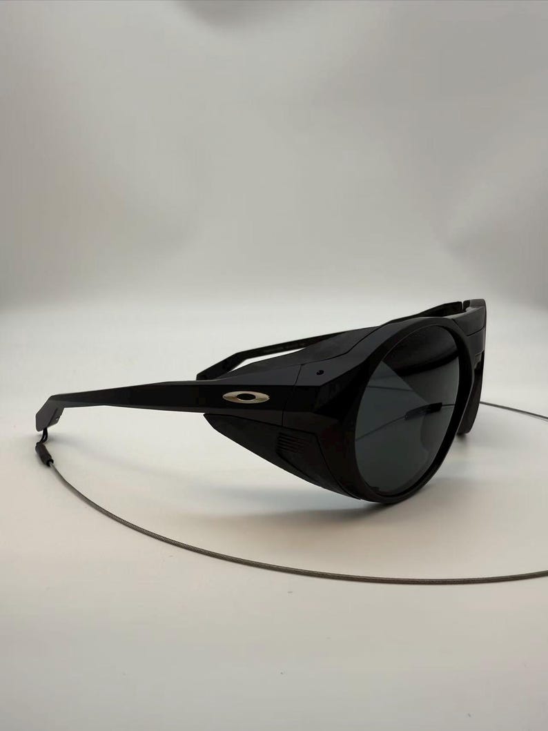 Puede incluir: Gafas de sol negras envolventes con lentes oscuras y montura negra. Las gafas de sol presentan un logotipo plateado en el lateral y un fino cord&oacute;n gris sujeto a la montura.