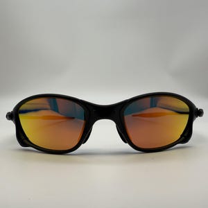 May include: Black sunglasses with orange and yellow mirrored lenses. The sunglasses have a sleek, modern design with a curved frame and a bridge over the nose. The lenses reflect a gradient of colors.