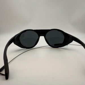 Puede incluir: Gafas de sol negras con un dise&ntilde;o envolvente &uacute;nico. Las lentes son oscuras y las monturas son de color negro mate. Las patillas son finas y se extienden hacia atr&aacute;s desde las lentes. Las gafas de sol parecen ser de pl&aacute;stico.
