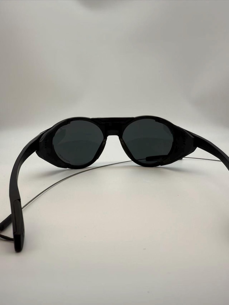 Puede incluir: Gafas de sol negras con un dise&ntilde;o envolvente &uacute;nico. Las lentes son oscuras y las monturas son de color negro mate. Las patillas son finas y se extienden hacia atr&aacute;s desde las lentes. Las gafas de sol parecen ser de pl&aacute;stico.
