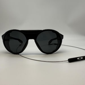 Puede incluir: Gafas de sol negras con lentes redondas y montura gruesa. Las gafas tienen un puente que conecta las lentes y una fina correa plateada con una pieza final negra. El logotipo de Oakley es visible en la correa.