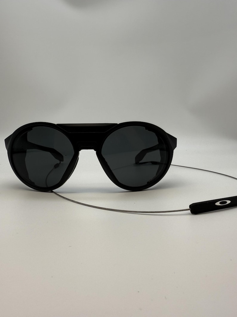 Puede incluir: Gafas de sol negras con lentes redondas y montura gruesa. Las gafas tienen un puente que conecta las lentes y una fina correa plateada con una pieza final negra. El logotipo de Oakley es visible en la correa.