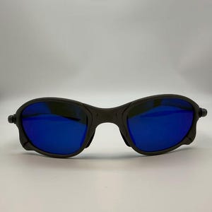 Puede incluir: Gafas de sol negras con lentes de espejo azules. Las gafas tienen una montura redondeada y un dise&ntilde;o deportivo. Las lentes reflejan un color azul brillante, y la montura es de color negro mate. Las gafas est&aacute;n dise&ntilde;adas para actividades al aire libre.