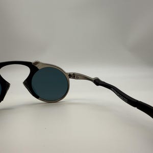 May include: Round sunglasses with a silver frame and dark blue lenses. The temples are black with the word "MANMAN" printed on the side. The sunglasses have a modern, minimalist design.