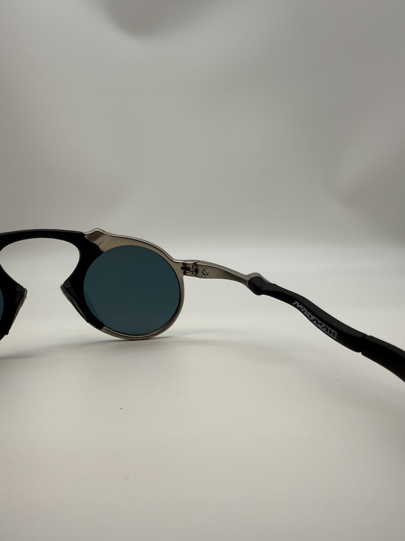 May include: Round sunglasses with a silver frame and dark blue lenses. The temples are black with the word "MANMAN" printed on the side. The sunglasses have a modern, minimalist design.