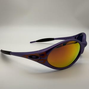 May include: Purple sunglasses with orange mirrored lenses. The frames have an orange and brown flame design. The arms have black tips. The sunglasses are set against a white background.