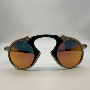 May include: Oval-shaped sunglasses with a silver frame and orange-tinted lenses. The sunglasses have a black bridge and a unique design. The lenses appear to have a polarized coating.