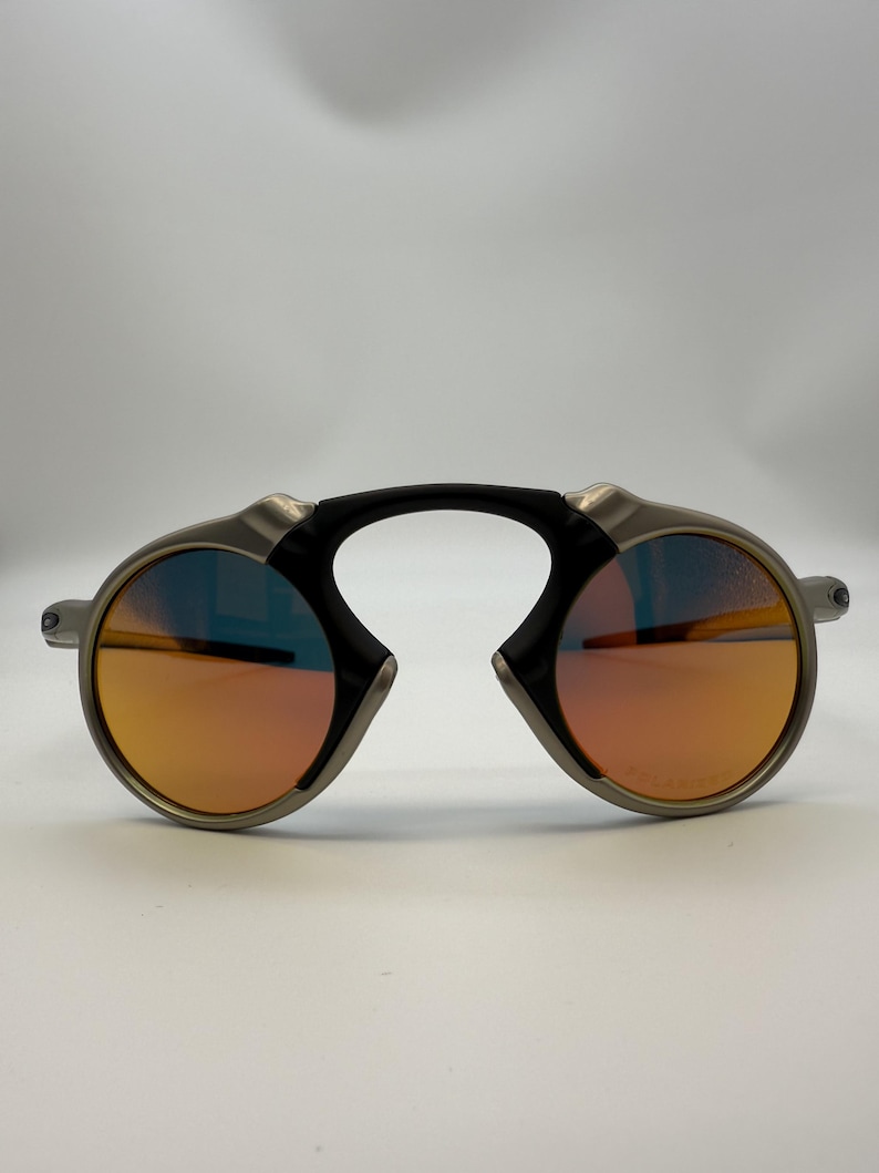 May include: Oval-shaped sunglasses with a silver frame and orange-tinted lenses. The sunglasses have a black bridge and a unique design. The lenses appear to have a polarized coating.