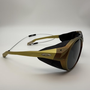 Könnte beinhalten: Goldene und schwarze Sonnenbrille mit dunklen Gläsern. Die Sonnenbrille hat einen goldenen Rahmen und Bügel, mit schwarzen Akzenten an den Seiten und oben. Die Gläser sind dunkel, und das Design ist sportlich.