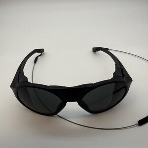 Puede incluir: Gafas de sol negras con lentes oscuras y un dise&ntilde;o envolvente. Las gafas de sol tienen un aspecto elegante y moderno y un fino cord&oacute;n plateado sujeto a las monturas. El cord&oacute;n tiene una pieza de goma negra en el extremo.