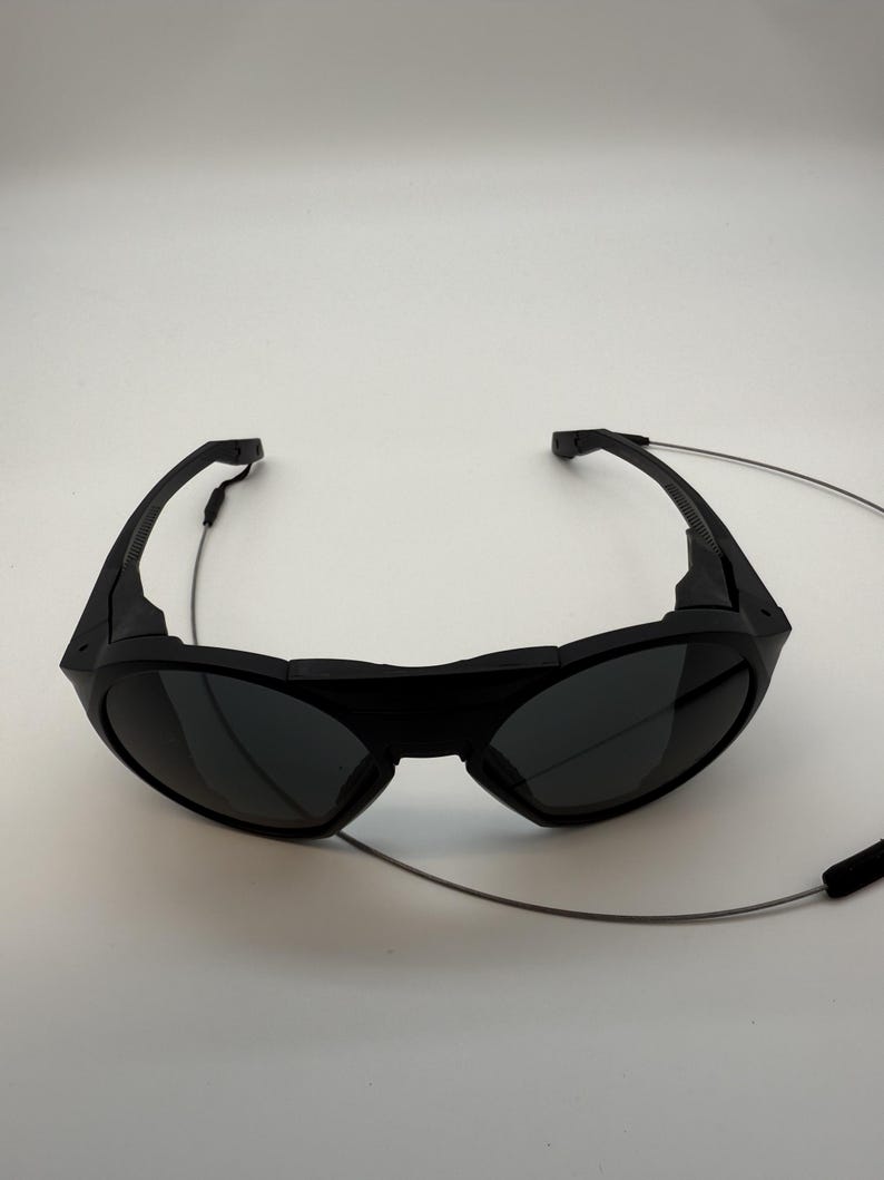 Puede incluir: Gafas de sol negras con lentes oscuras y un dise&ntilde;o envolvente. Las gafas de sol tienen un aspecto elegante y moderno y un fino cord&oacute;n plateado sujeto a las monturas. El cord&oacute;n tiene una pieza de goma negra en el extremo.