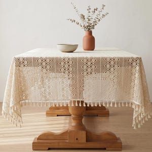 May include: Cream-colored lace tablecloth with a decorative border and tassel trim. The tablecloth is draped over a wooden table, with a vase of dried flowers and a bowl on top. A rustic, elegant home decor item.