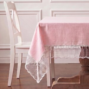 May include: A pink tablecloth with a white lace trim and a white chair. The tablecloth is draped over a wooden table. The lace trim has a scalloped edge. The chair has a white finish and a cross-back design.