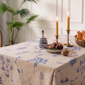 May include: A beige tablecloth with a blue floral pattern covers a table set for a meal. Two lit candles in brass holders, a plate of figs, and a decorative bell are on the table. A basket of bread is also visible.