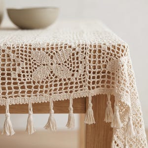 Peut inclure: Nappe en crochet crème avec un motif floral et des glands en franges. La nappe est drapée sur une table en bois, mettant en valeur le motif ajouré complexe et le bord décoratif.