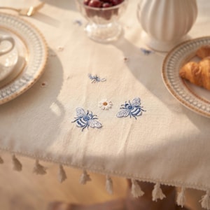 Puede incluir: Mantel crema con abejas azules bordadas y una margarita. La mesa está puesta con platos con bordes dorados, una jarra blanca y un cuenco de cristal con cerezas. Borlas adornan el borde del mantel.