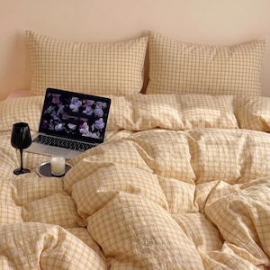 Op de afbeelding: Een bed met gele geruite beddengoed, inclusief kussens en een dekbed. Een laptop met een bloemrijk scherm wordt op het bed weergegeven, samen met een zwart wijnglas en een kleine kaars op een dienblad. De algehele esthetiek is gezellig en uitnodigend.