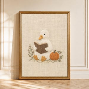 May include: A framed embroidery art piece on a textured beige fabric background featuring a white duck with an orange beak and feet, holding an open brown book, accompanied by a small orange pumpkin and green foliage accents.