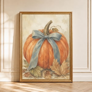 May include: A framed wall art print featuring a large orange pumpkin with a prominent light blue ribbon tied around its stem. The pumpkin rests on a bed of dried autumn leaves against a neutral, textured background, all encased in a gold frame.