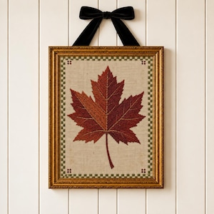 May include: A framed embroidery piece featuring a large, detailed maple leaf in shades of deep red, orange, and brown. The leaf is centered on a light beige fabric with a green and white checkered border. The frame is gold, topped with a black velvet bow.