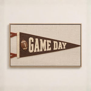 Puede incluir: Un cuadro decorativo que muestra un banderín de fieltro marrón con el texto GAME DAY en letras de bloque color crema. Un icono de balón de fútbol americano marrón y blanco se sitúa a la izquierda del texto. El banderín tiene borde crema.