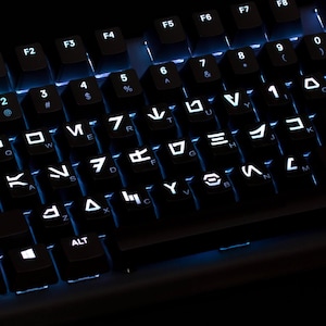 Könnte beinhalten: Eine schwarze Computertastatur mit hintergrundbeleuchteten Tasten und weißen Aurebesh-Zeichen. Die Tasten sind in einem Standardlayout angeordnet, mit Funktionstasten oben und einem blauen Lichtschein unter den Tastenkappen.