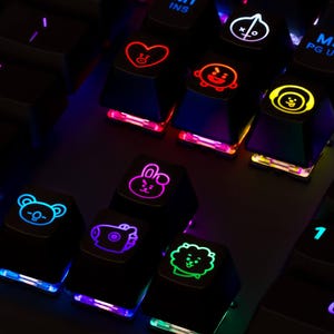 May include: A black mechanical computer keyboard featuring backlit keys with colorful character icons. The keys display glowing symbols including a red heart, a yellow character with headphones, a pink rabbit, and a green lion-like figure.