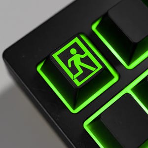 May include: Close-up of a black keyboard with green illuminated keys. The focus is on the exit key, which features a green graphic of a person running through a door. The key is square and surrounded by a green glow.