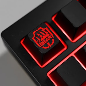 May include: Close-up of a black keyboard with red backlighting. The keycaps are black, with a red graphic of a bear claw and the word "BEAR" on one key. The keyboard is designed for gaming.